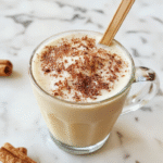 A close-up of a steaming vanilla cinnamon latte in a clear glass mug, topped with whipped cream and a cinnamon stick, with a sprinkle of cinnamon and vanilla bean pods on a rustic wooden table, warm lighting accentuates the creamy texture and inviting aroma.